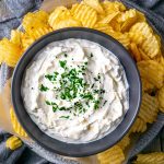 French onion dip in black bowl over platter of potato chips on gray linen on gray wood table