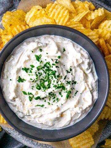 French onion dip in black bowl over platter of potato chips on gray linen on gray wood table