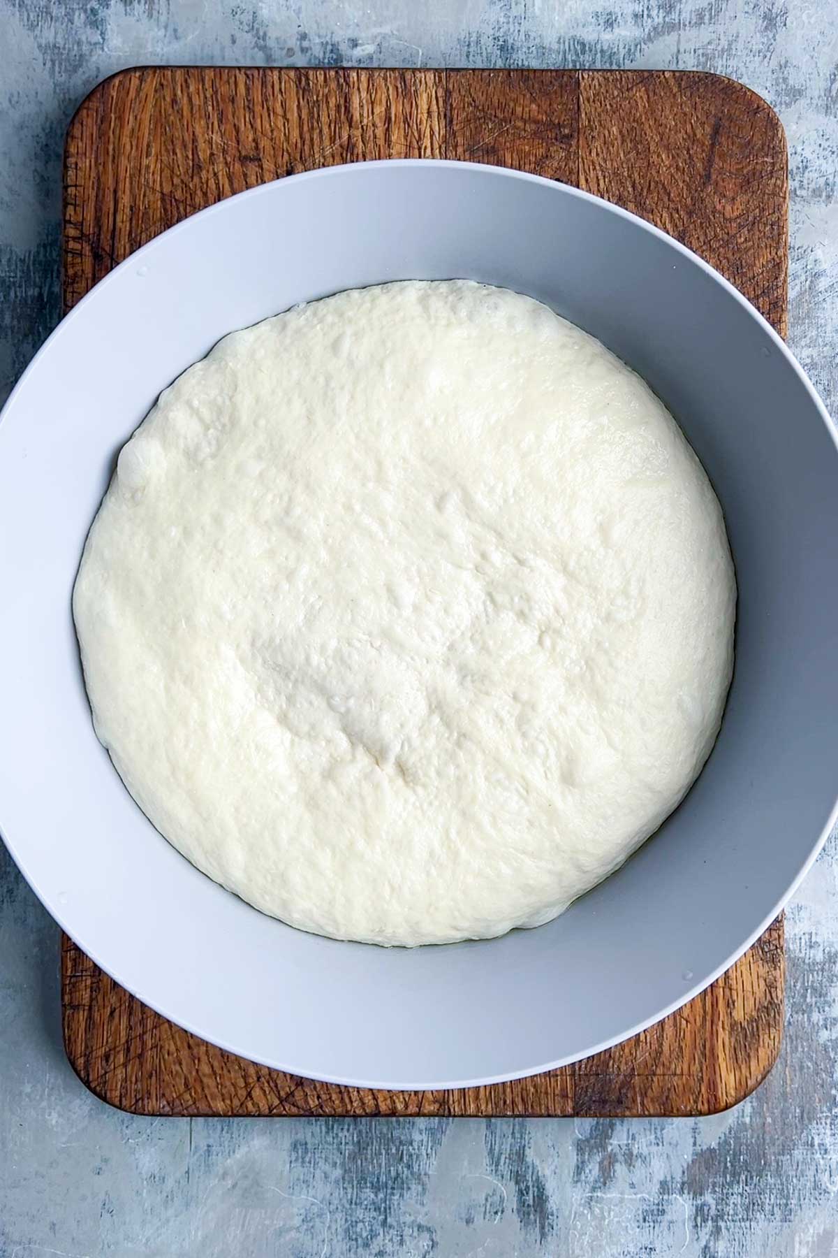 A bowl of risen naan bread dough sits on a wooden cutting board against a light grey textured background.