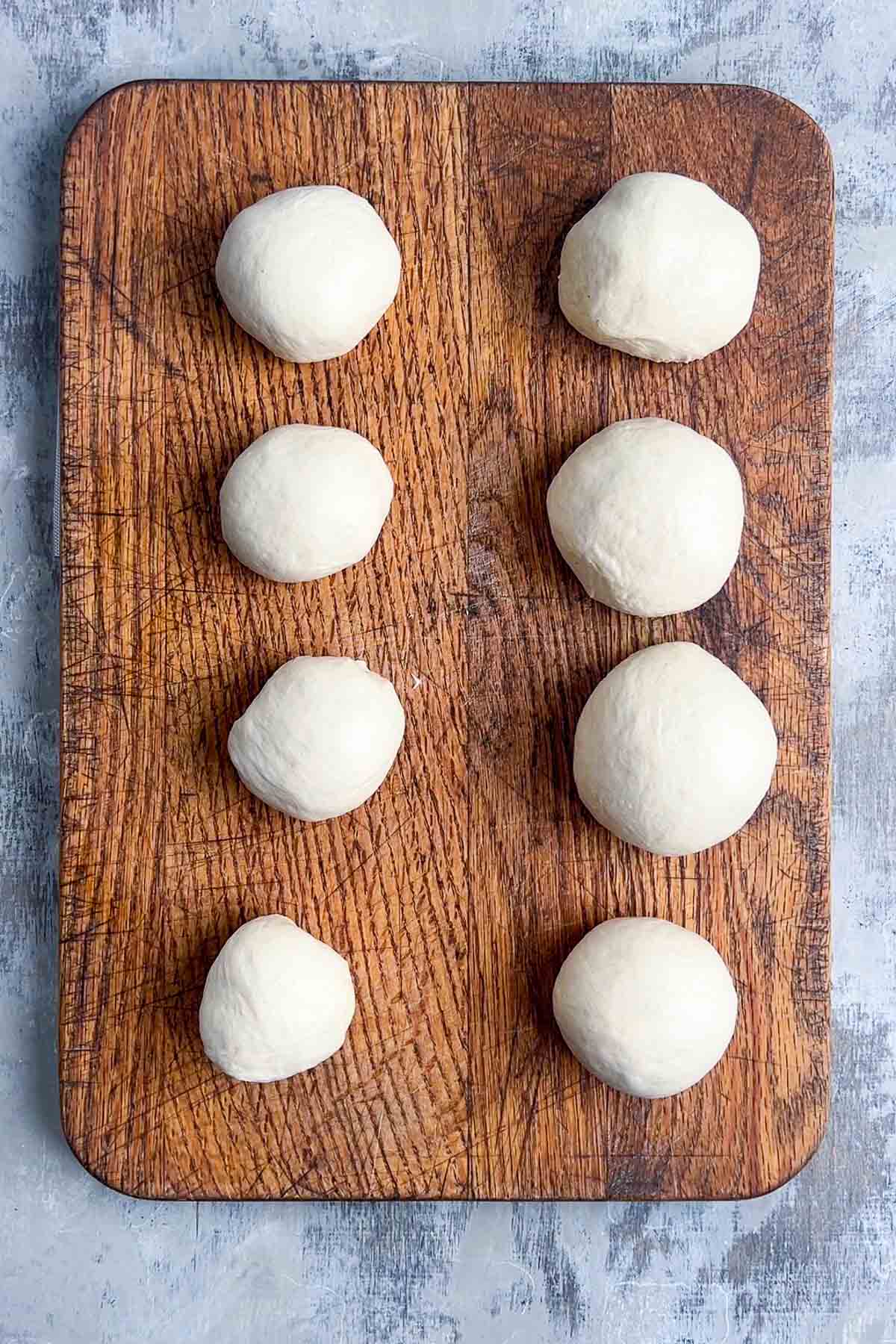 Eight balls of bread dough arranged in two columns on a wooden cutting board, viewed from above.