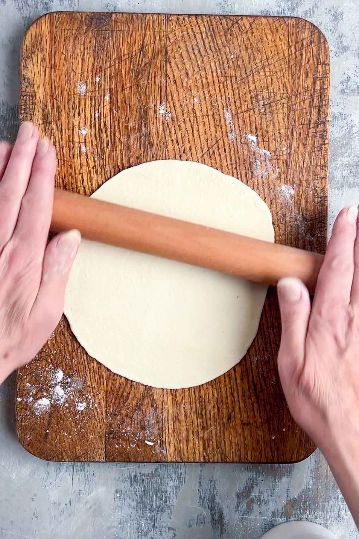 Two hands use a rolling pin to flatten dough for naan bread on a wooden cutting board, with some flour scattered on the board and surface.