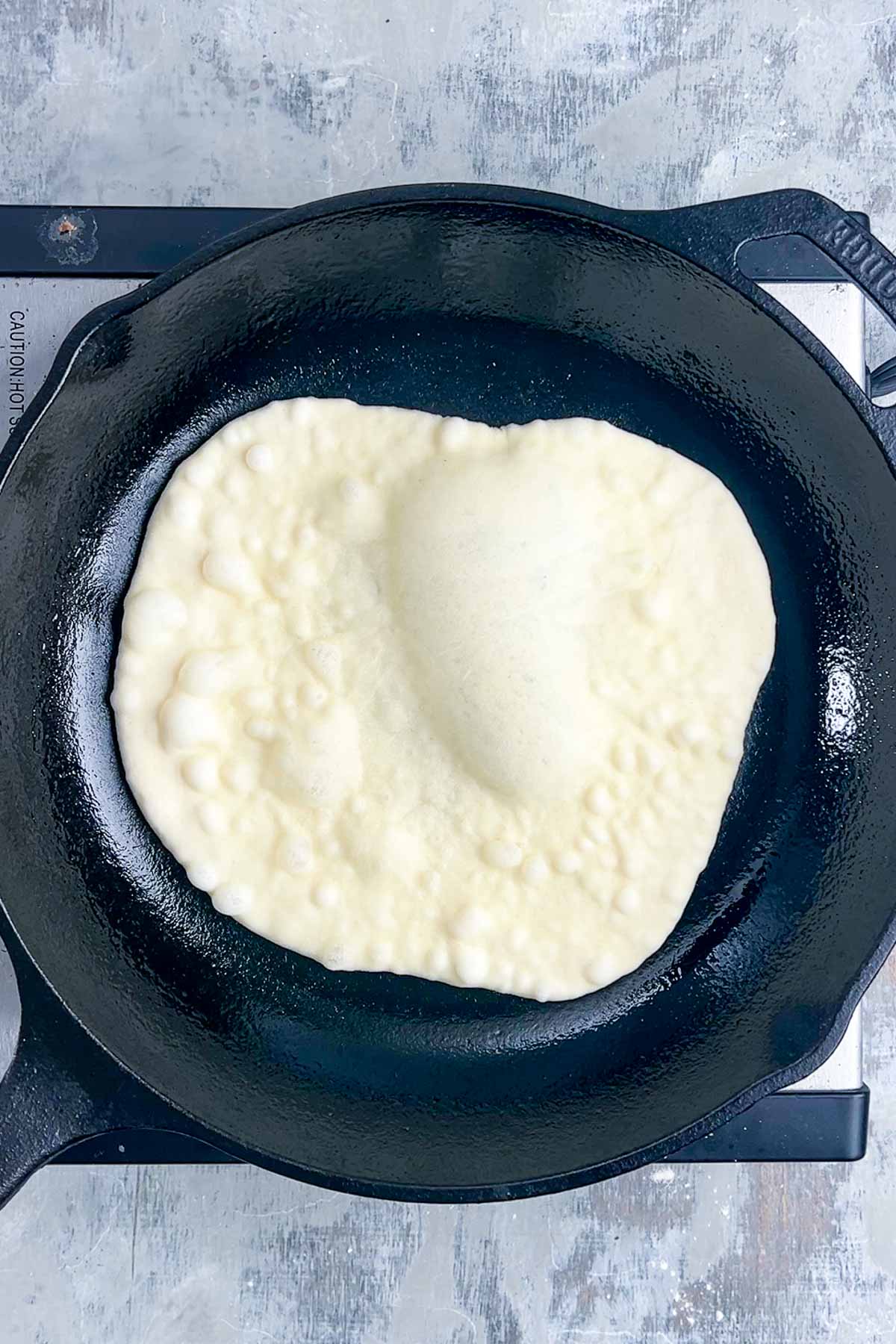 A piece of naan bread dough cooking in a black cast iron skillet on a stovetop, with bubbles forming on the surface.