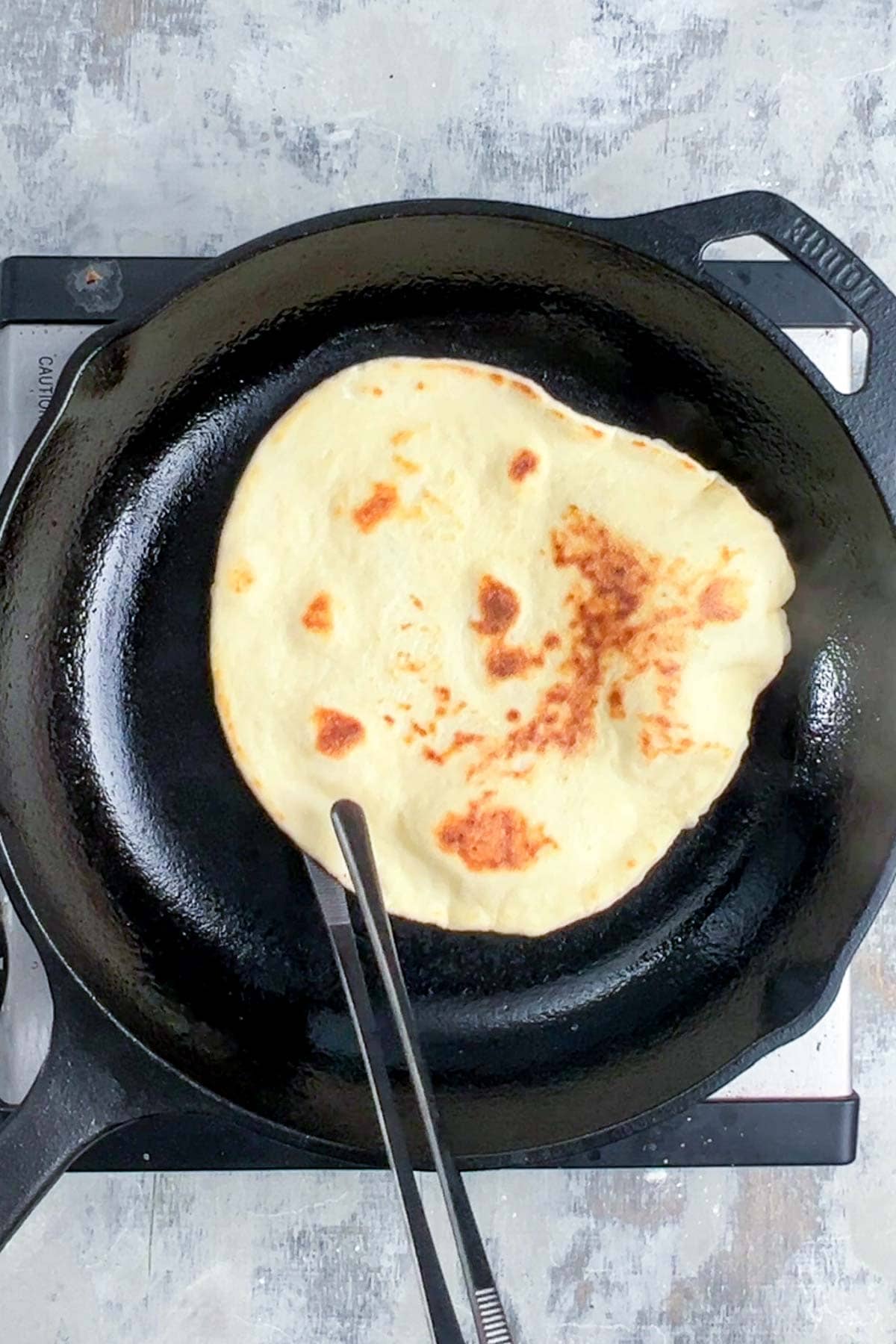 A piece of naan bread is being cooked in a black cast iron skillet on a stovetop, with tongs holding one side of the bread.