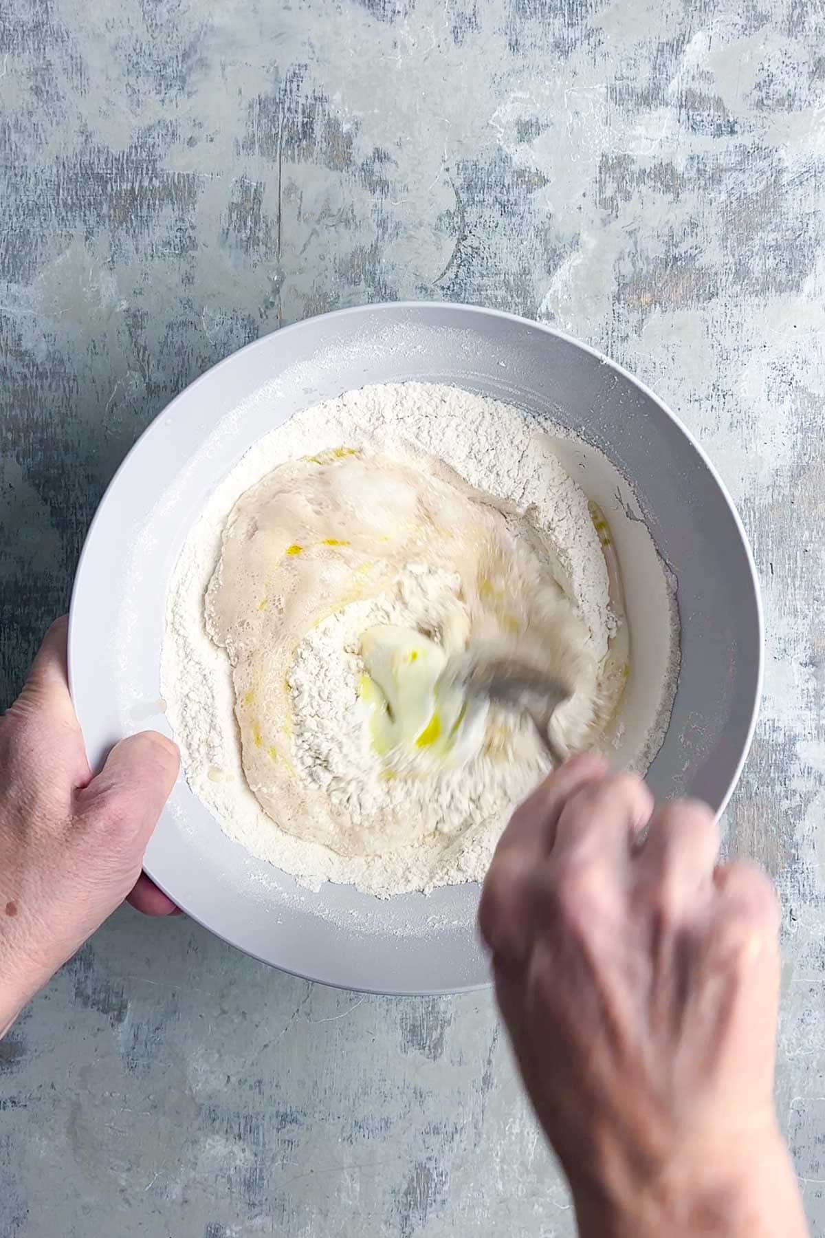 Two hands mix flour, yogurt, water, and oil in a white bowl with a fork for naan bread.