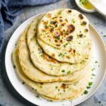 A plate of stacked buttery garlic naan bread garnished with fresh parsley, with a small bowl of melted butter and a pastry brush beside it.