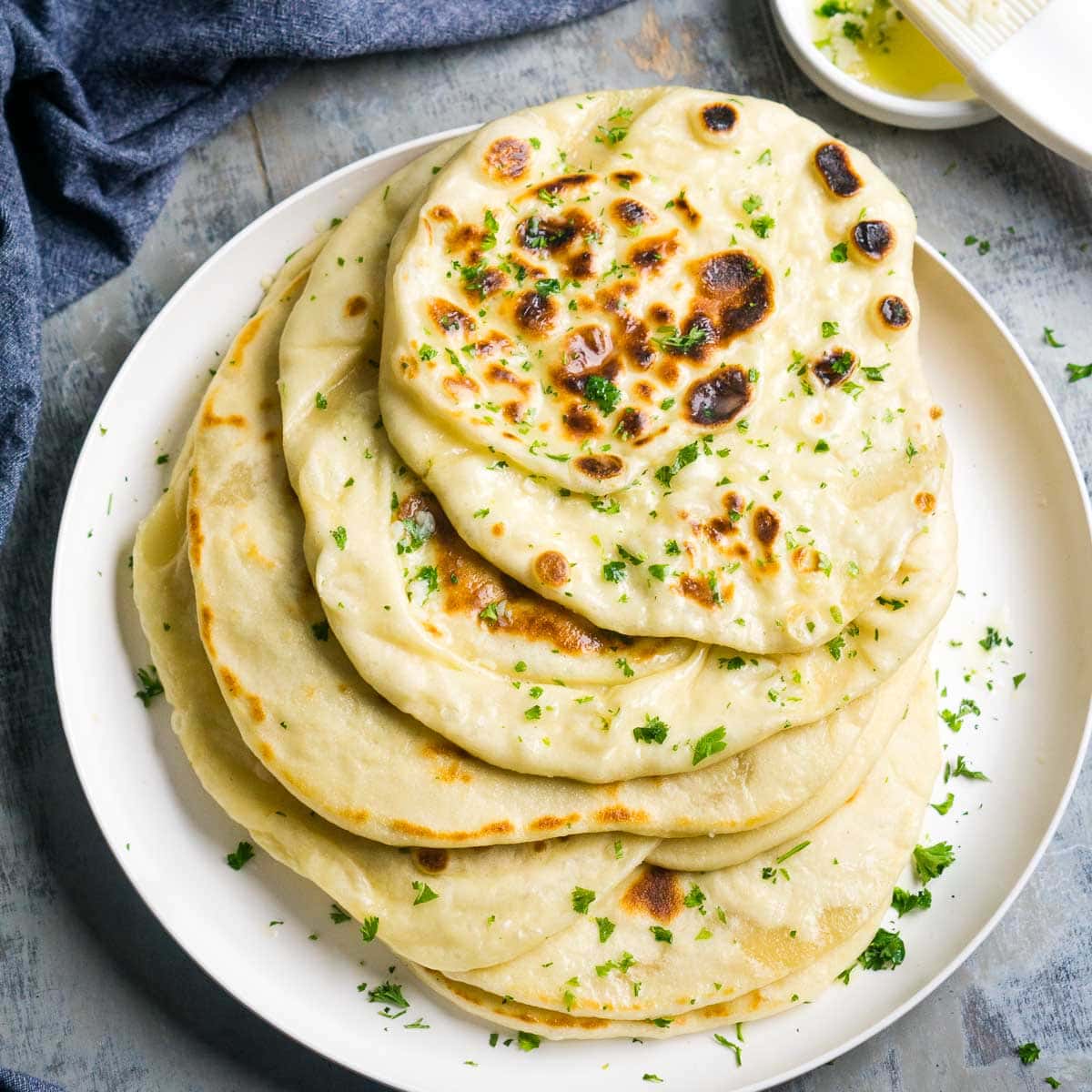 A plate of stacked buttery garlic naan bread garnished with fresh parsley, with a small bowl of melted butter and a pastry brush beside it.