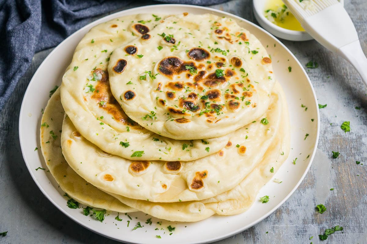 A plate of stacked buttery garlic naan bread garnished with fresh parsley, with a small bowl of melted butter and a pastry brush beside it.