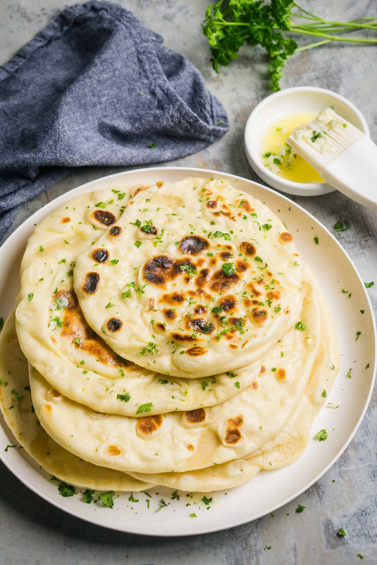 A plate of stacked buttery garlic naan bread garnished with fresh parsley, with a small bowl of melted butter and a pastry brush beside it.