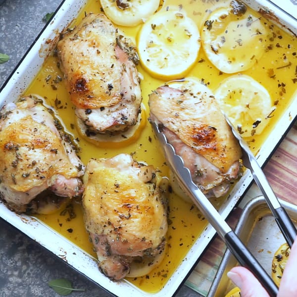 tongs moving Greek lemon chicken thighs from baking sheet to black serving plate