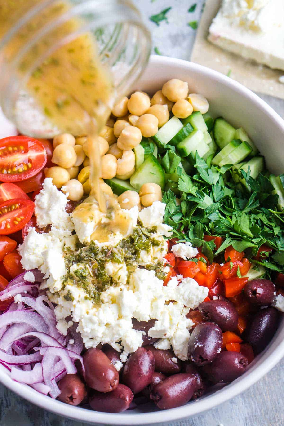 Greek dressing is pouring over Greek chickpea salad in white bowl