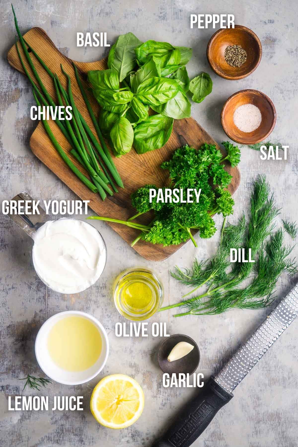 Chives, basil, parsley, dill, Greek yogurt, olive oil, garlic, lemon juice, salt, and pepper arranged on a surface and labeled&mdash;classic ingredients for a vibrant green goddess dressing.