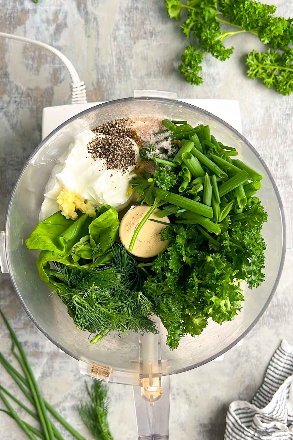 A food processor bowl with Greek yogurt, garlic, black pepper, salt, parsley, dill, chives, and basil&mdash;ready to make green goddess dressing. Fresh herbs and a striped towel are nearby.