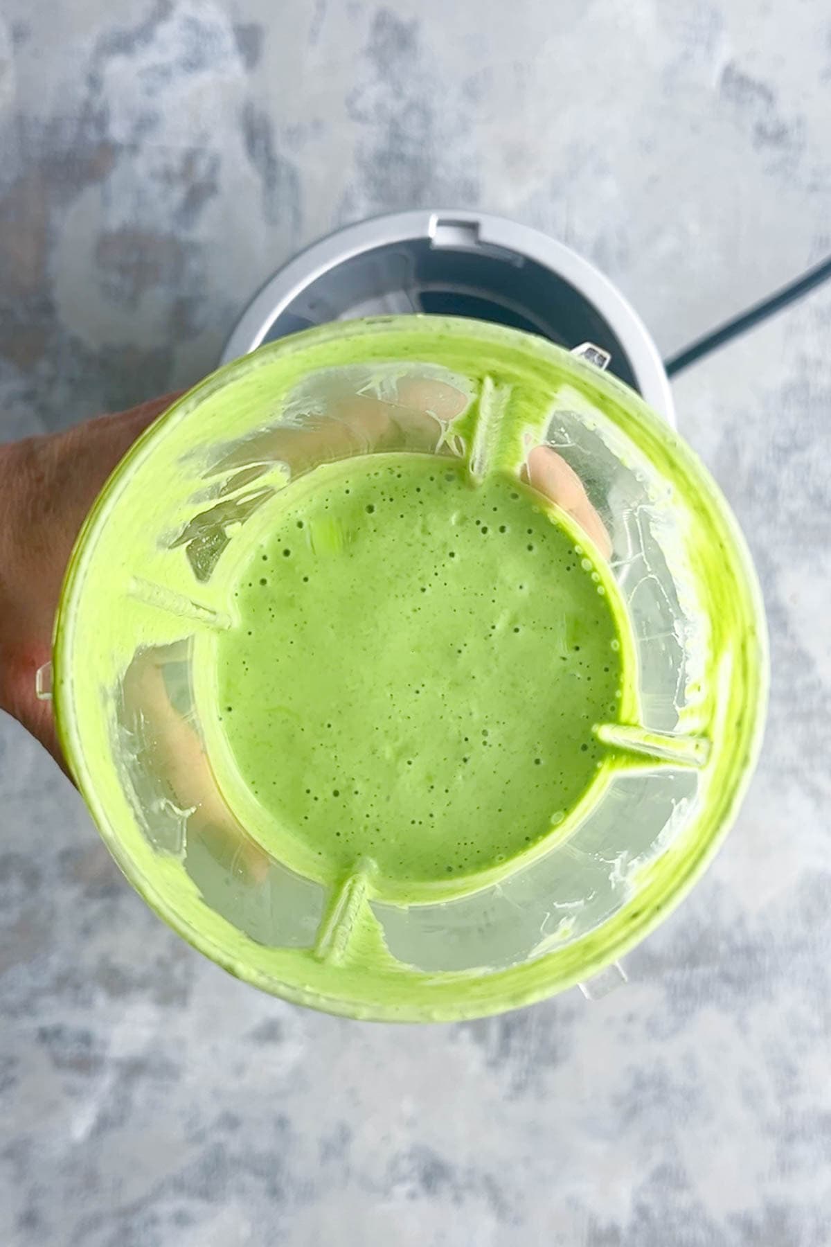 A hand holds a high speed blender cup filled with freshly blended green goddess dressing, viewed from above against a light gray textured background.
