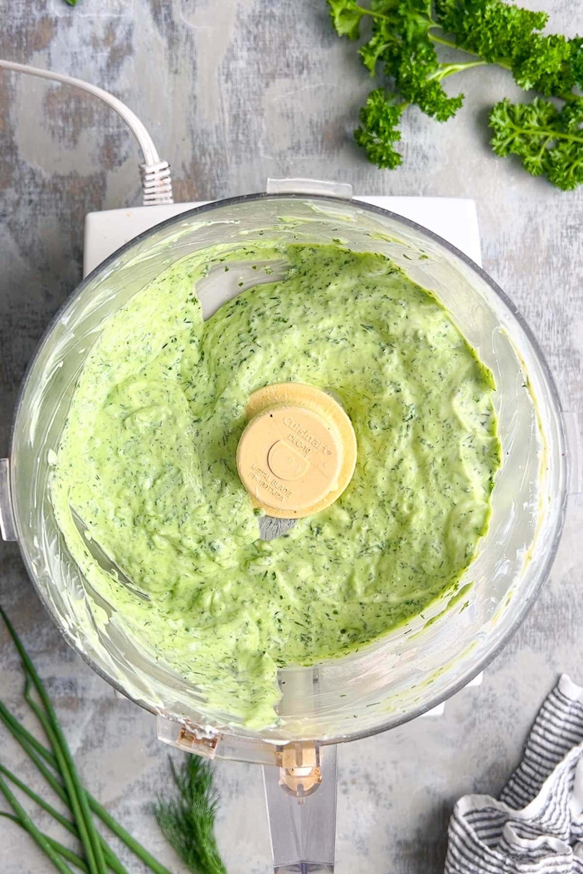 Food processor containing a creamy green goddess dressing on a counter, with fresh parsley, dill, and chives nearby.