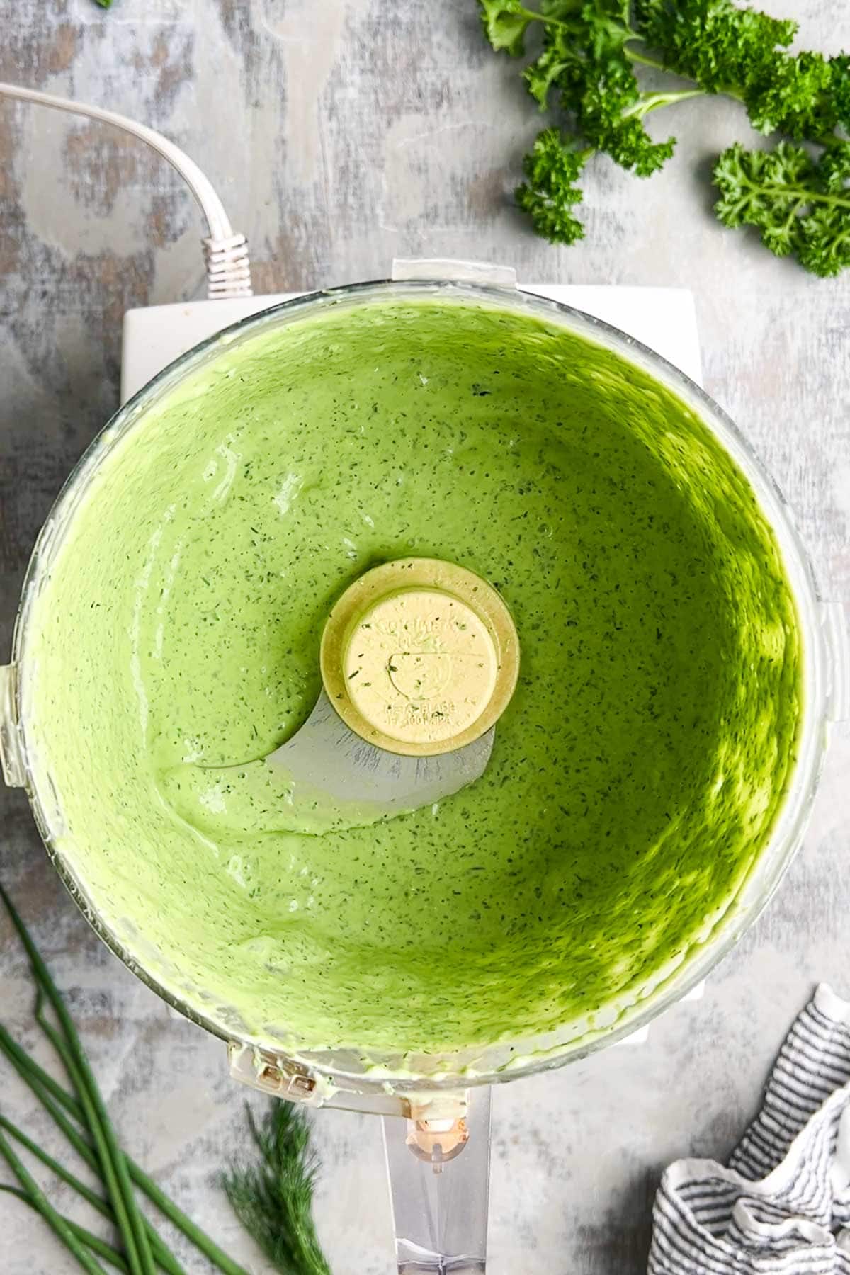 A food processor bowl containing vibrant green goddess dressing sits on a counter, with fresh parsley, chives, and dill nearby.