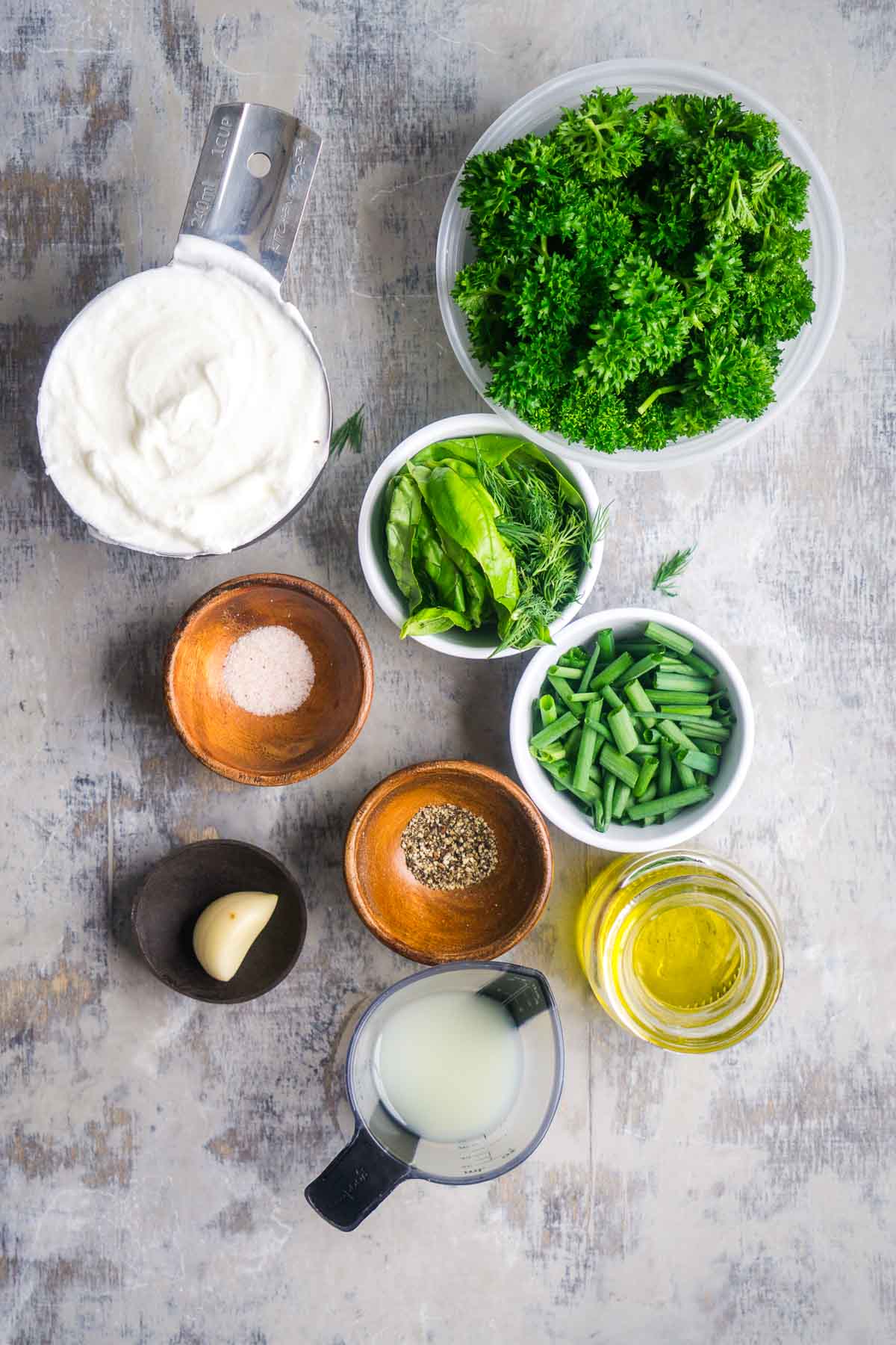 Assorted ingredients on a gray surface, including plain yogurt, parsley, basil, dill, chives, salt, pepper, garlic, olive oil, and a small cup of lemon juice&mdash;perfect for making homemade green goddess dressing.