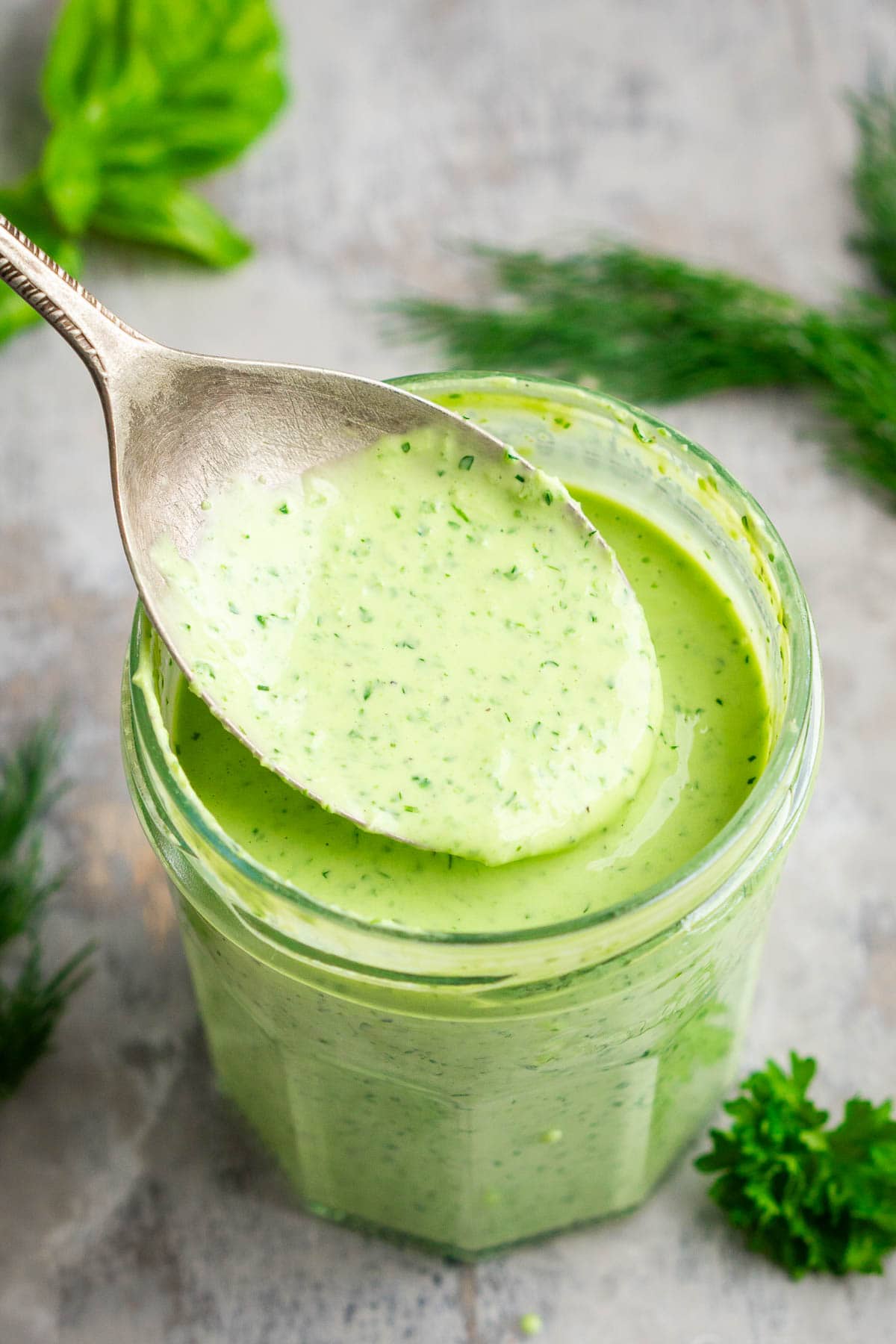 A metal spoon holds creamy green goddess dressing above a glass jar full of the dressing, surrounded by fresh herbs on a gray surface.