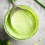 Overhead closeup of a spoon holding creamy green goddess dressing above a glass jar filled with the dressing; fresh herbs are scattered nearby.