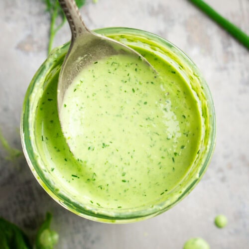 Overhead closeup of a spoon holding creamy green goddess dressing above a glass jar filled with the dressing; fresh herbs are scattered nearby.
