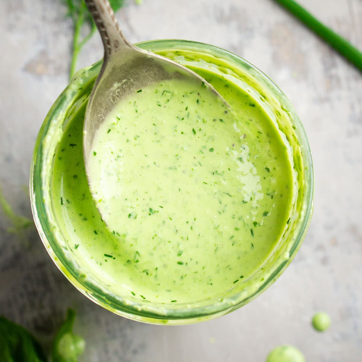 Overhead closeup of a spoon holding creamy green goddess dressing above a glass jar filled with the dressing; fresh herbs are scattered nearby.
