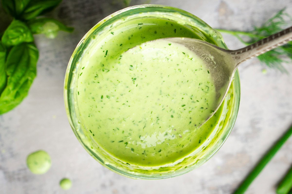 A bowl of creamy green goddess dressing with a spoon, photographed from above. Fresh herbs are visible in the vibrant mixture and scattered on the table nearby.