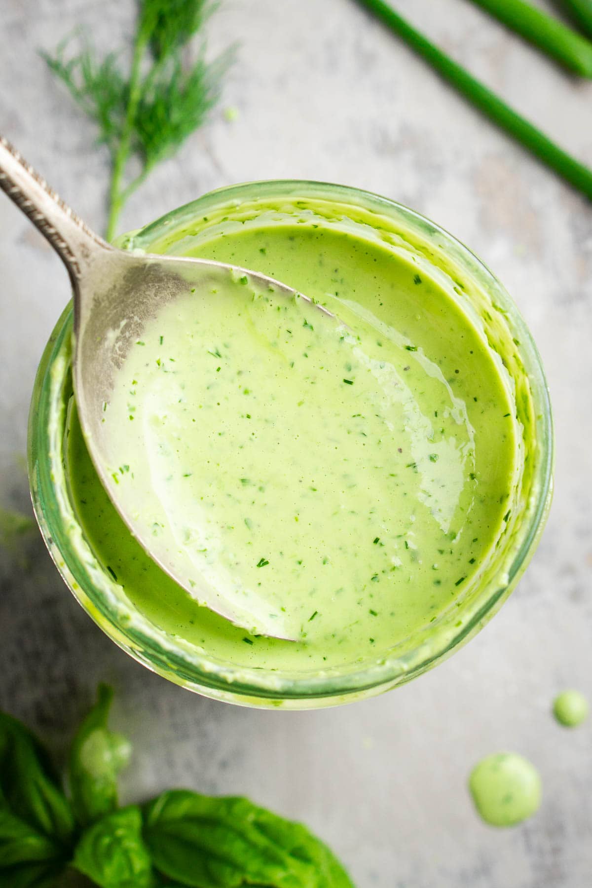Overhead closeup of a spoon holding creamy green goddess dressing above a glass jar filled with the dressing; fresh herbs are scattered nearby.
