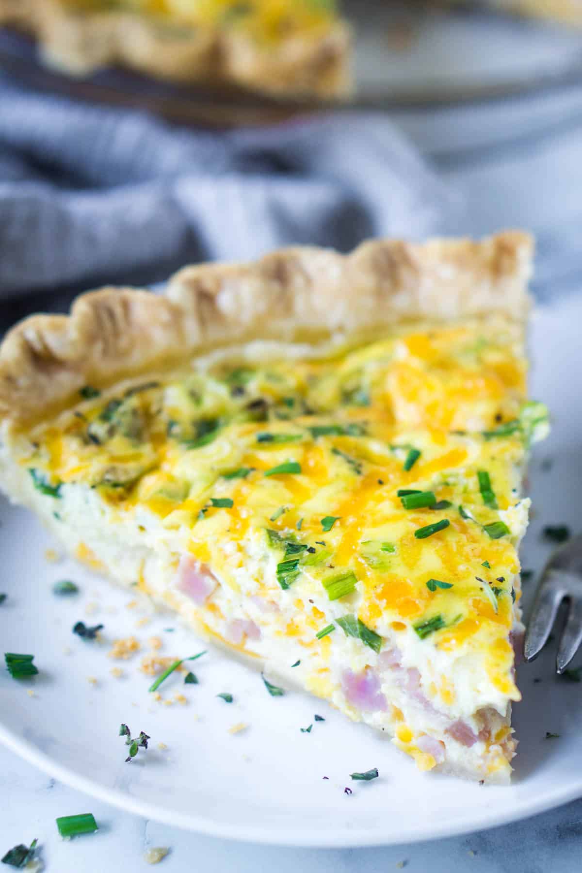 Slice of ham and cheese quiche on plate with fork next to linen.