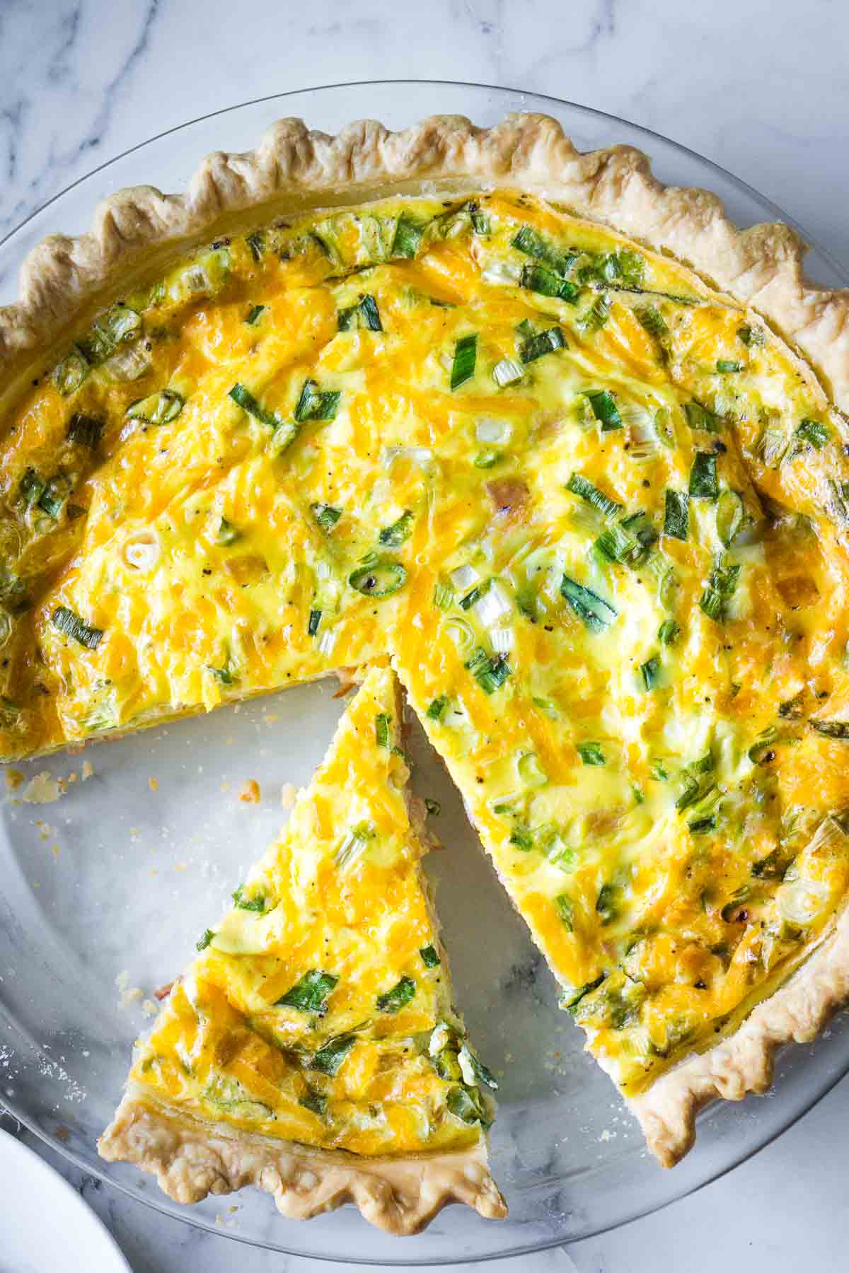 A sliced quiche with a golden crust, filled with ham and cheese, green onions, and vegetables, sits enticingly on a clear glass pie dish.
