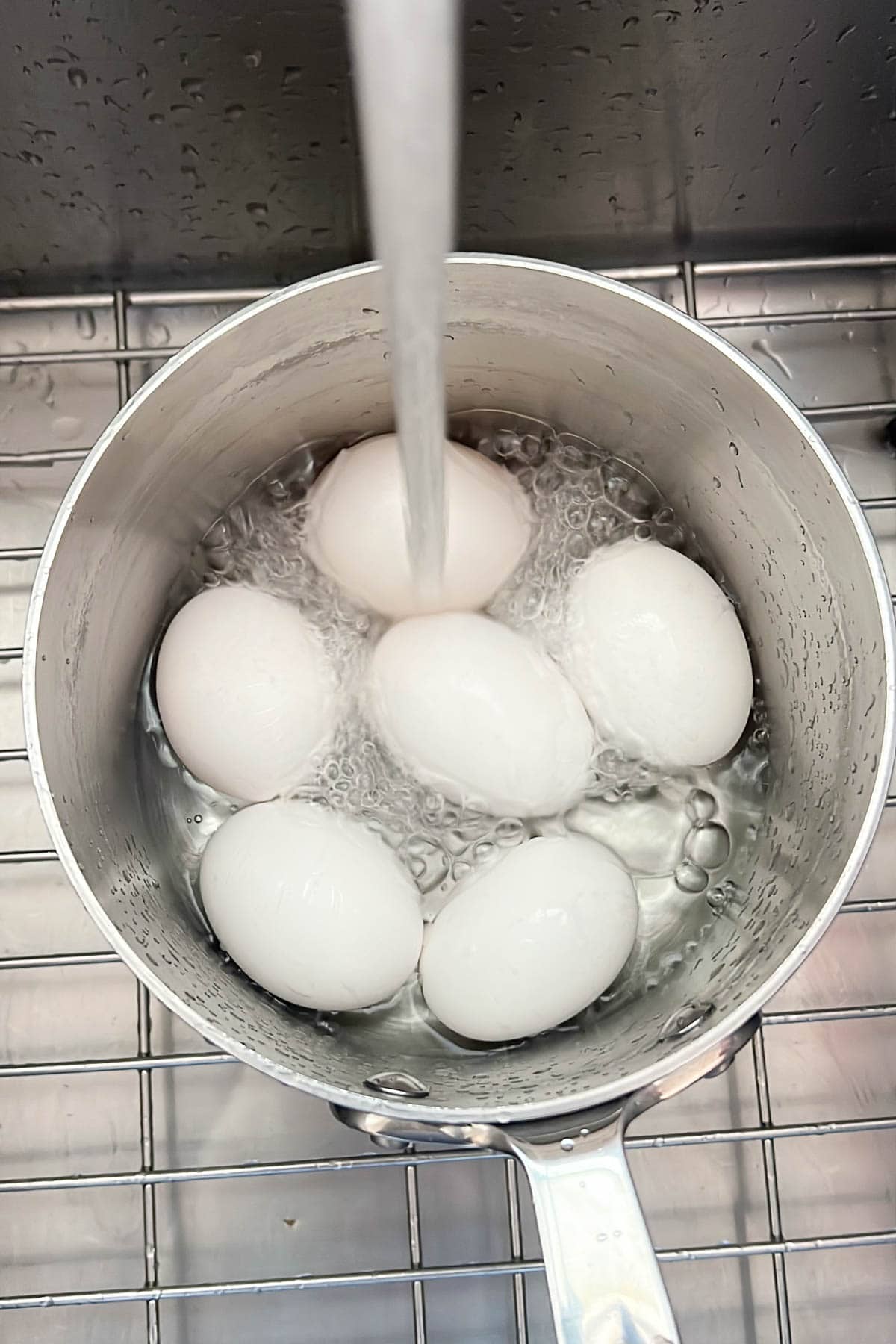 Six hard boiled eggs are being cooled under running water in a metal pot in a kitchen sink, ensuring they&rsquo;re perfectly cooked and easy to peel.