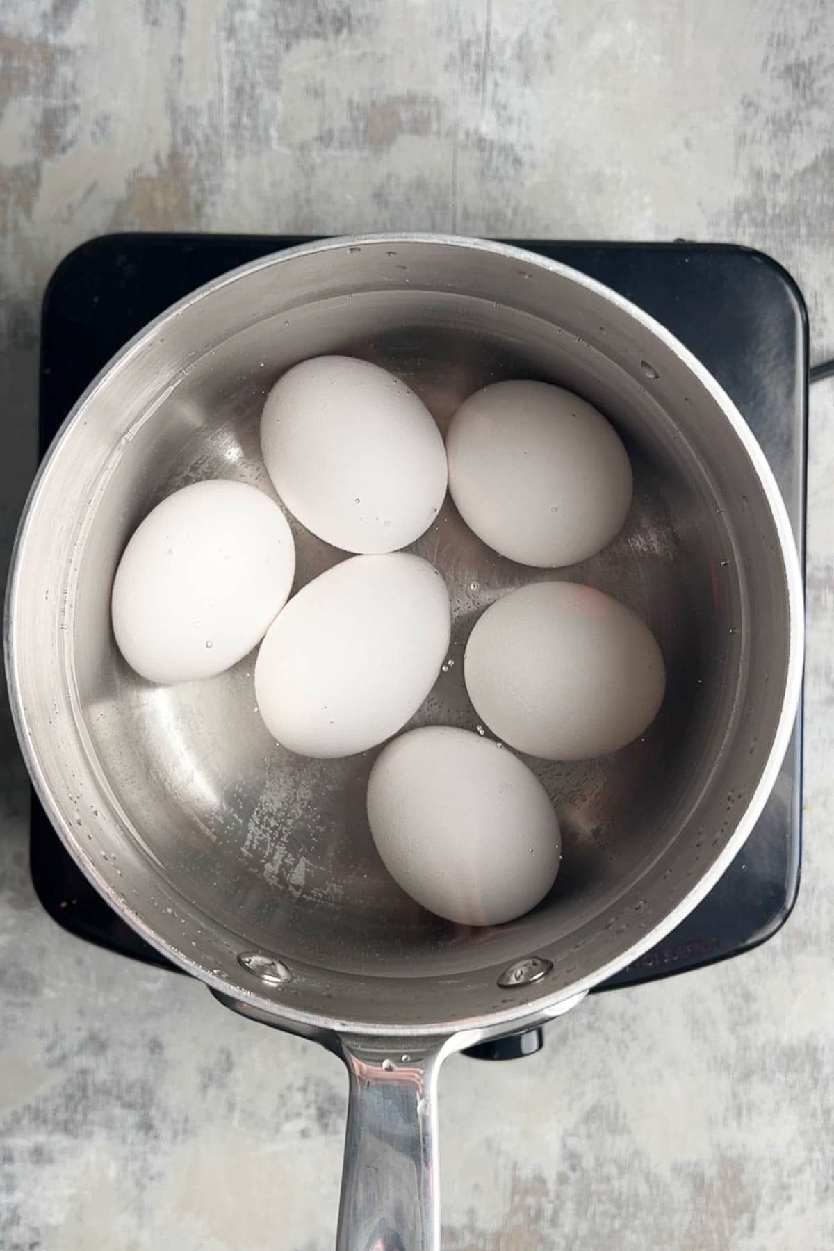 Six white eggs in a pot of water on a stovetop, ready to be cooked into perfect hard boiled eggs.