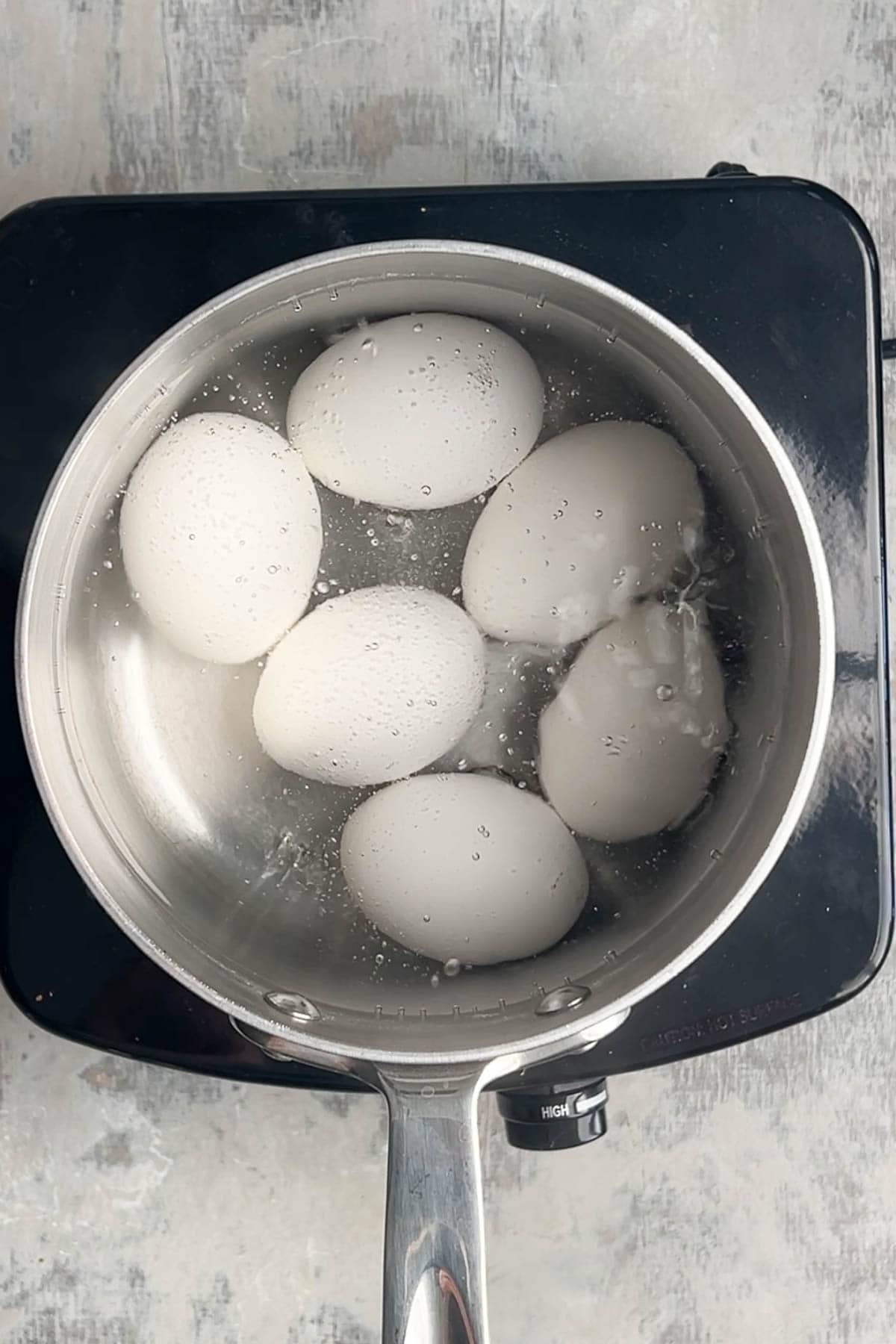 Six hard boiled eggs are being cooked in water at a rolling boil in a stainless steel pot on an electric stovetop.