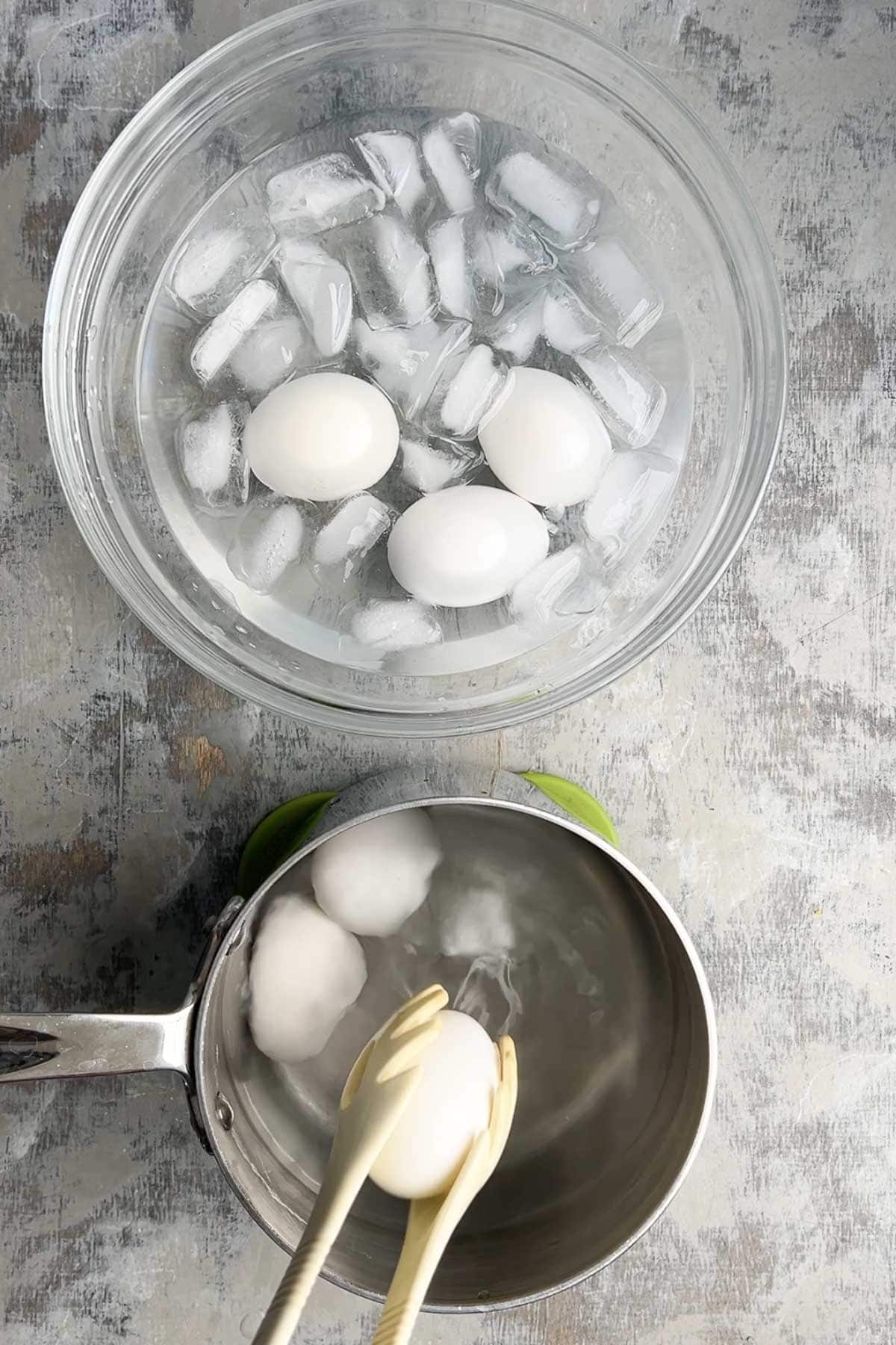 A person uses tongs to transfer hard boiled eggs from a pot of hot water to a bowl of ice water.