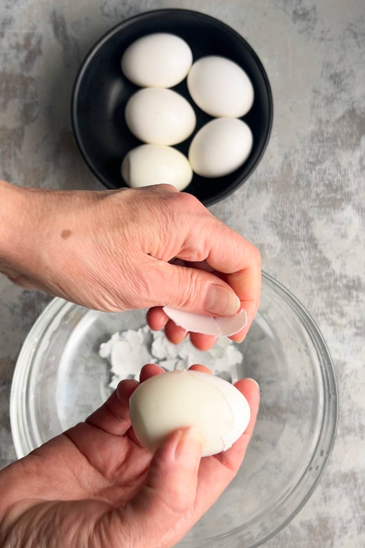 Two hands peel hard boiled eggs over a clear bowl filled with eggshell pieces, while a black bowl with more unpeeled hard boiled eggs rests on the gray surface nearby.