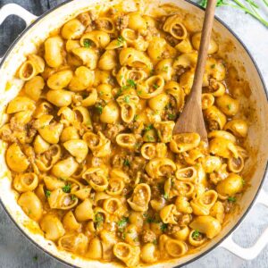 A skillet filled with creamy pasta shells, ground meat, and garnished with chopped herbs. A wooden spoon is resting in the skillet.