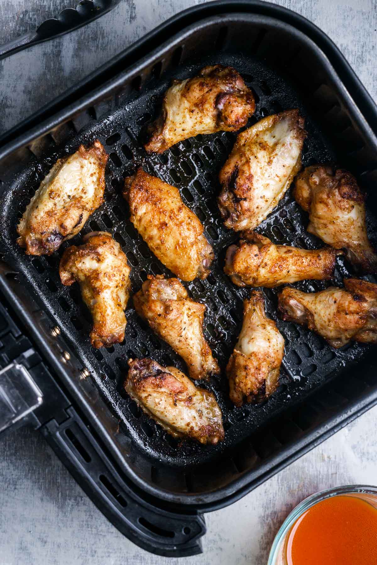 crispy chicken wings in square air fryer basket next to hot wing sauce