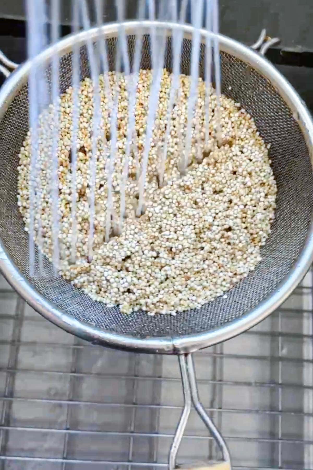 Water is being poured over uncooked quinoa in a fine mesh strainer positioned above a sink.