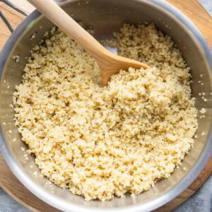A stainless steel pot filled with cooked quinoa being stirred with a wooden spoon, placed on a wooden cutting board.