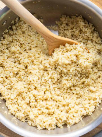 A stainless steel pot filled with cooked quinoa being stirred with a wooden spoon, placed on a wooden cutting board.