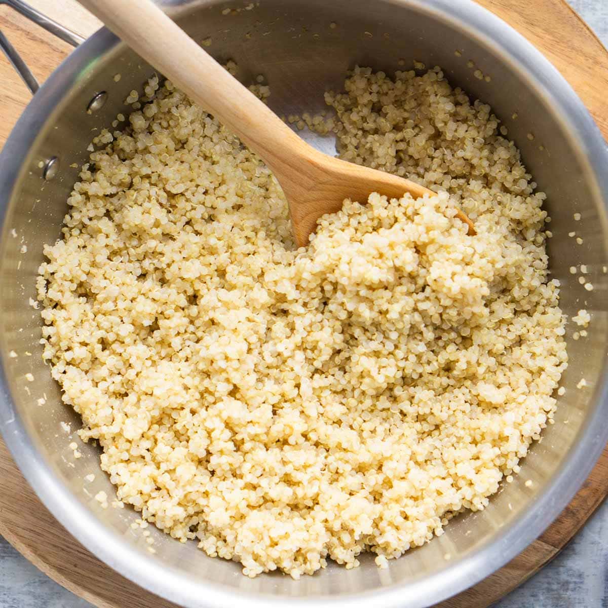 A stainless steel pot filled with cooked quinoa being stirred with a wooden spoon, placed on a wooden cutting board.