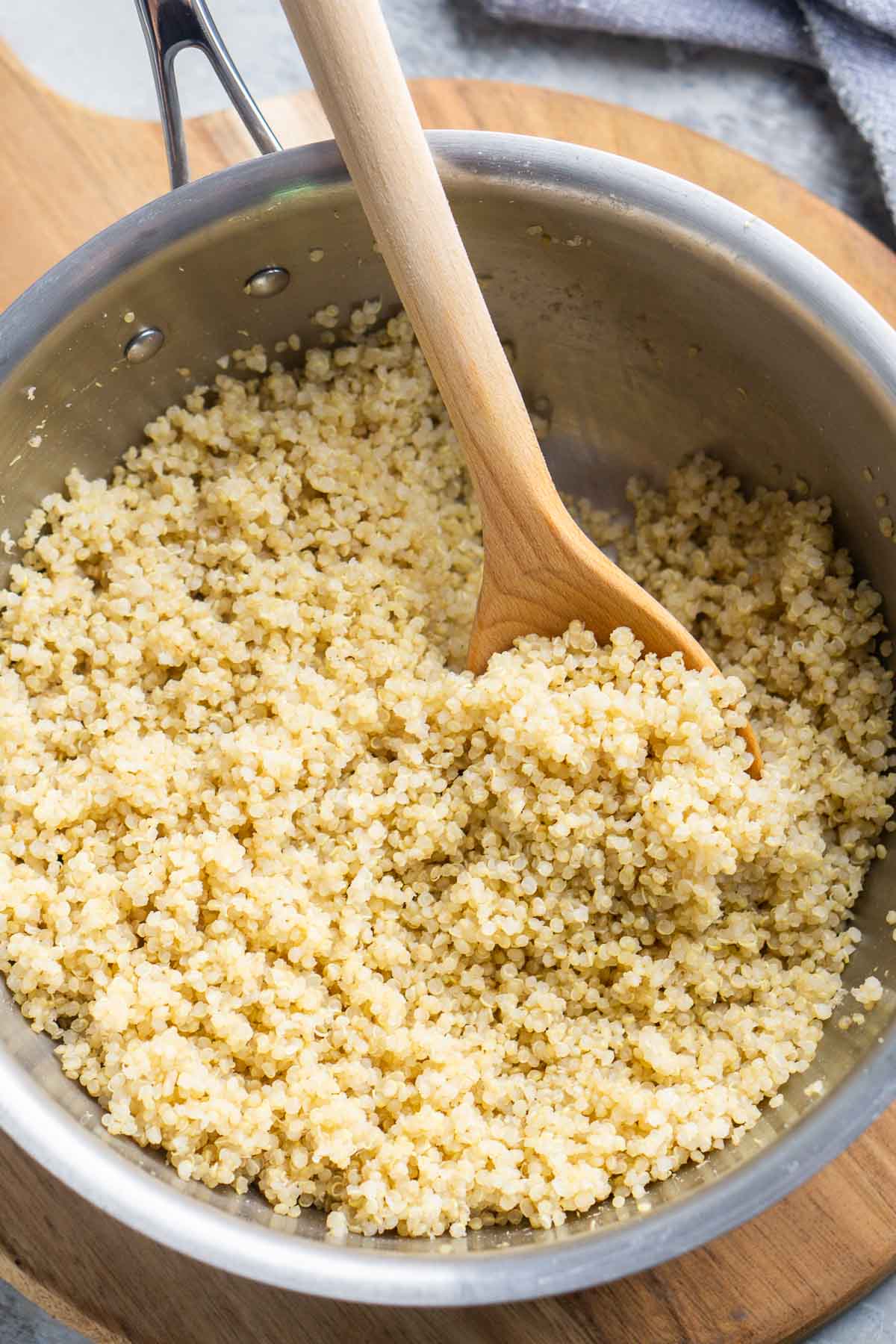 Learn how to cook quinoa on the stove with this simple, reliable method for fluffy quinoa every time. This recipe uses a proven quinoa-to-water ratio and works with white, red, and tri-color quinoa. A stainless steel pot filled with cooked quinoa being stirred with a wooden spoon, placed on a wooden cutting board next to a gray cloth.