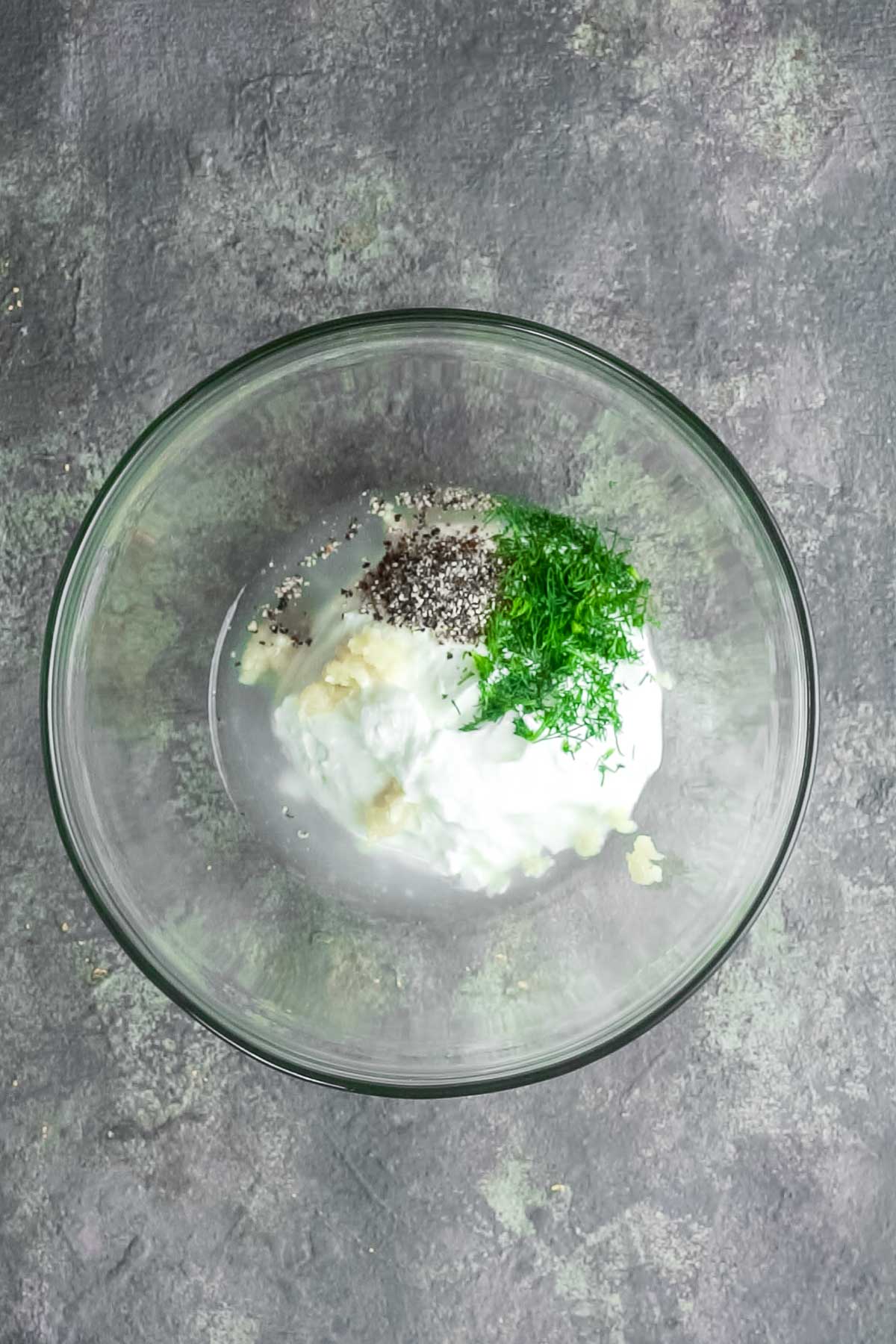 A glass bowl containing sour cream, chopped dill, minced garlic, black pepper, and salt on a gray textured surface.