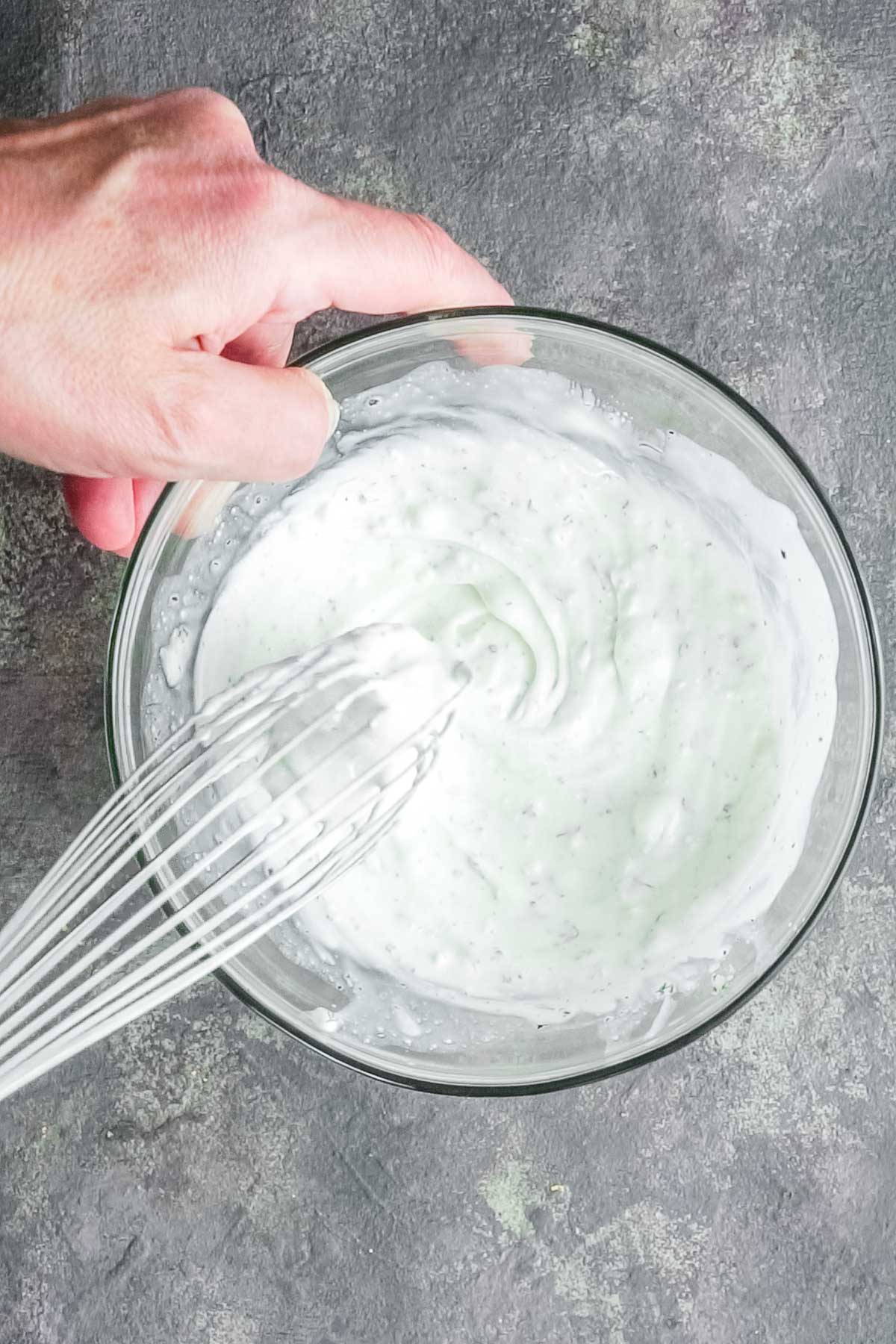 A hand holds a glass bowl while whisking a creamy white dressing.