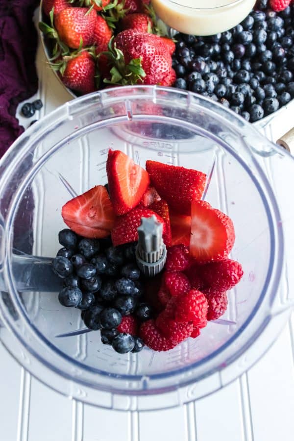 Strawberries, blueberries, and raspberries in food processor bowl next to bowl of fruit on white surface