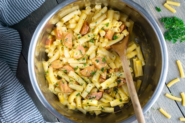 Wooden spoon stirring instant pot mac & cheese with smoked sausage. Pot is on gray background with blue and white striped linen, dry pasta noodles, and fresh parsley