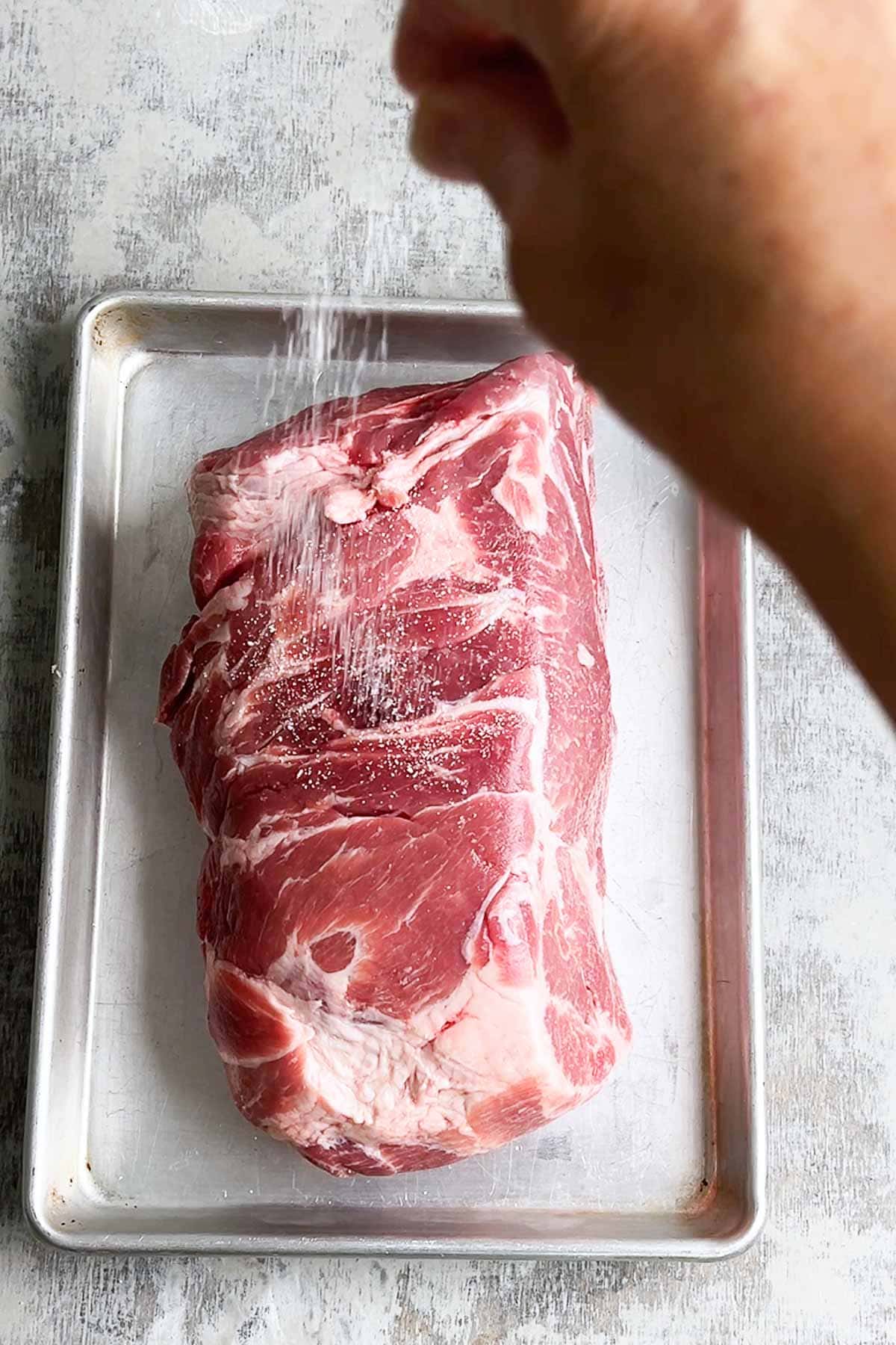 Person's hand sprinkles salt over pork butt on baking sheet.