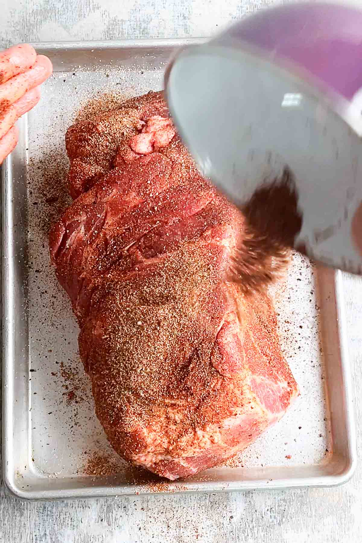 Person's hand sprinkles seasoning over pork butt on baking sheet.