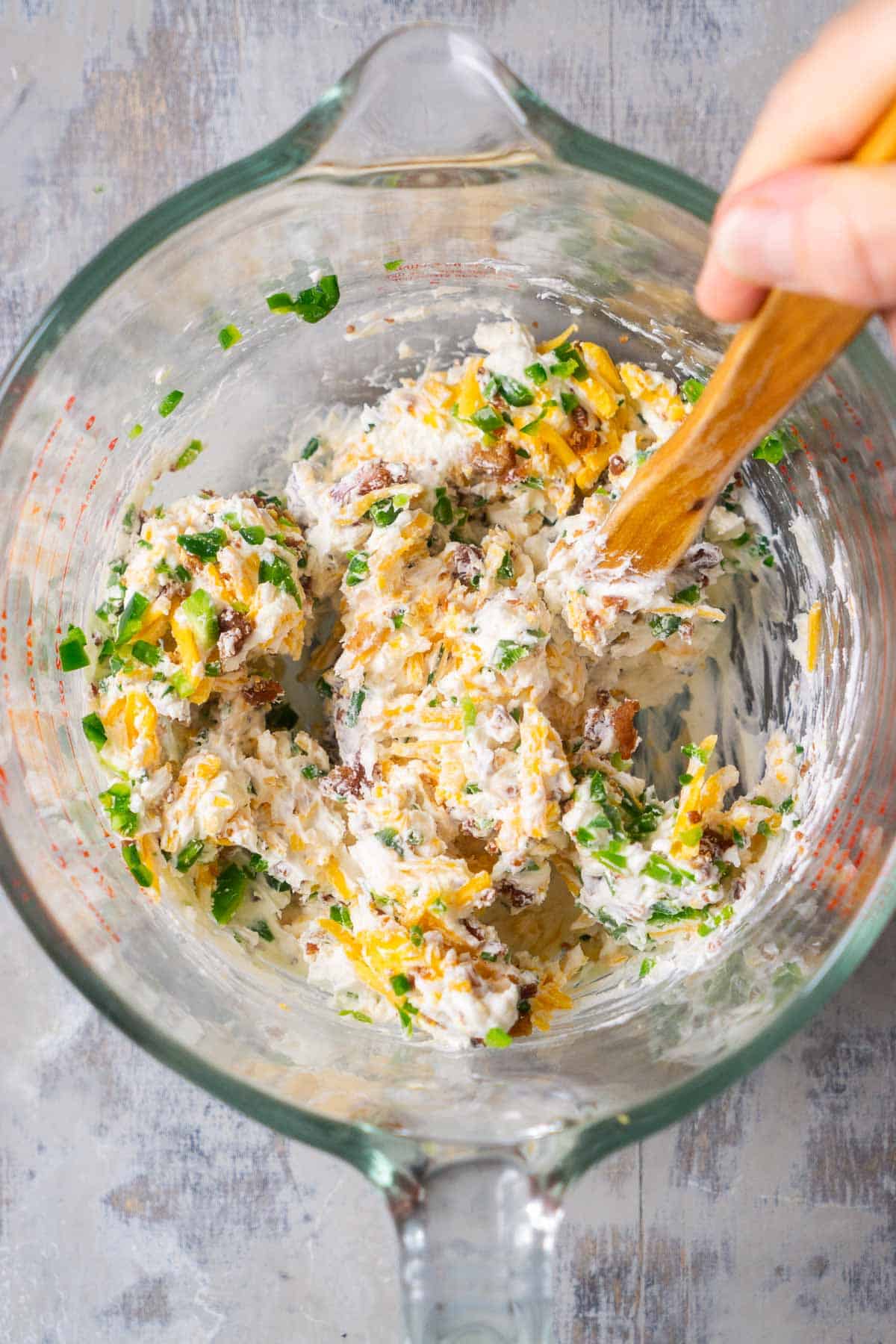 person's hand stirs jalapeno popper cheese ball ingredients together in glass mixing bowl