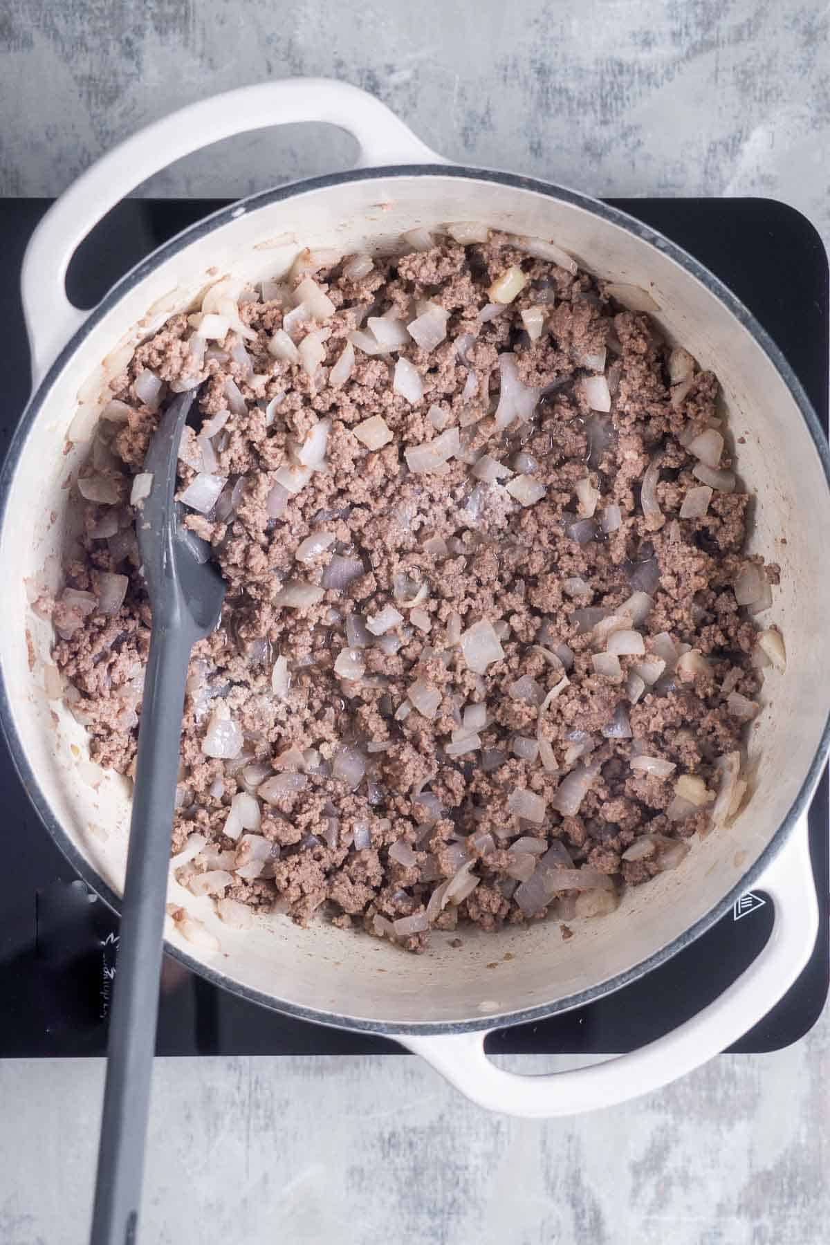 Gray spoon stirs cooked ground beef and onions in Dutch oven