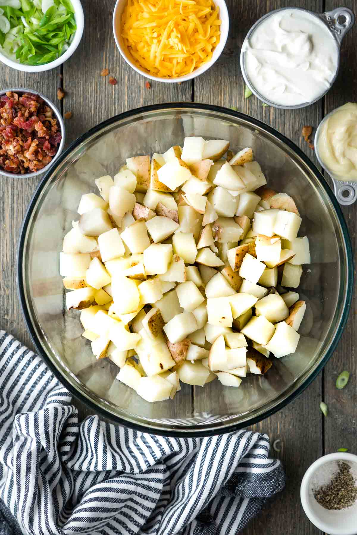 cooked diced potatoes in glass bowl next to potato salad ingredients in small bowls