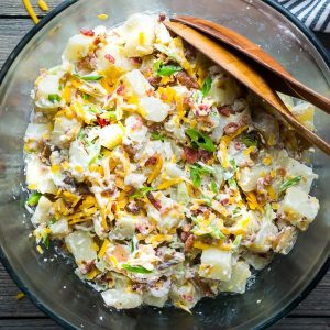 Loaded potato salad in large glass mixing bowl with wooden salad tongs on gray wood surface